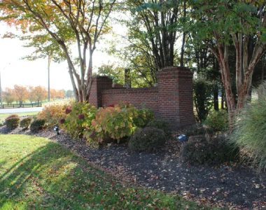 Brick sign with autumn foliage, trees, and shrubs on a landscaped area by a roadside.